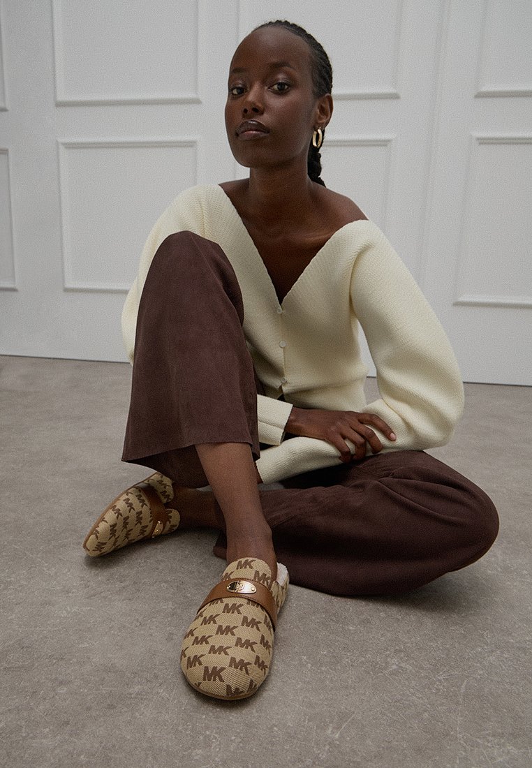 MK-mönstrade slip-on skor i brunt och beige, tillverkade av tyg, med ett läderrem och guldfärgade detaljer. Bärs med en krämfärgad tröja och bruna byxor.
