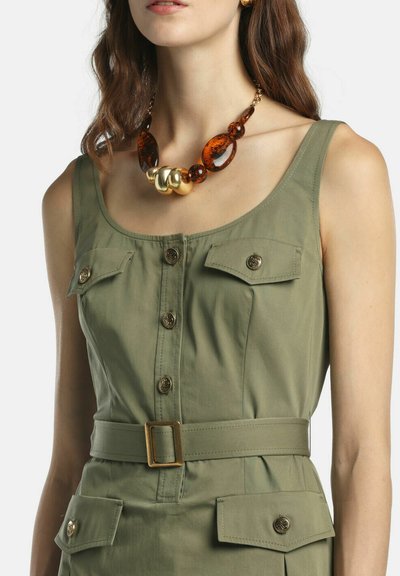 Sieviete valkā bezapkaklīti olīvu zaļas krāsas kleitu ar jostu, pogām aizpogātām kabatām un biezām dzintara un zelta krāsas krellēm.