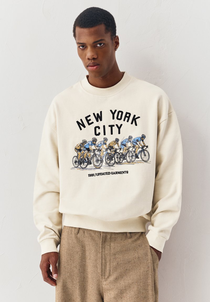 Jeune homme portant un pull beige avec le texte New York City et un graphique de cyclisme, assorti à un pantalon marron, debout contre un mur blanc.