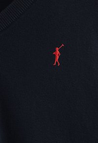 Granatowy sweter z dekoltem w serek, z małym czerwonym haftowanym logo przedstawiającym postać trzymającą kij golfowy po lewej stronie.