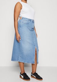 Ljusblå denimmaxikjol med en framficka, prydd med fickor, kombinerad med en vit ärmlös topp och svarta sneakers.