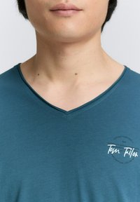 Teal V-kaula t-paita, joka on valmistettu pehmeästä kankaasta, jossa on pyöreä logoelementti vasemmalla puolella ja sileä pinta, jossa on ompeluyksityiskohtia.