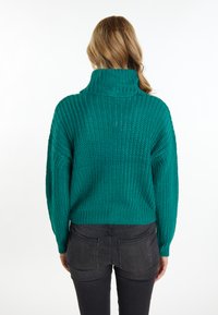 Pulover tricotat de culoare teal, cu guler înalt, având o croială lejeră și textură ribotată. Partea din spate are un design scurt, combinat cu blugi negri.