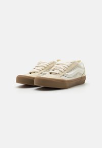 Beige suede sneakers with white accents, flat laces, and a brown rubber sole. Features a smooth texture and subtle logo detailing.