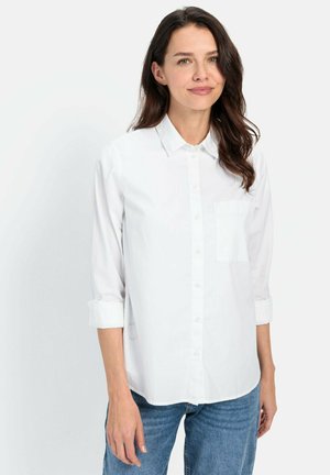 Chemise blanche à boutons avec une poche, manches longues roulées jusqu'au milieu de l'avant-bras, fabriquée en tissu léger, portée avec un jean bleu.