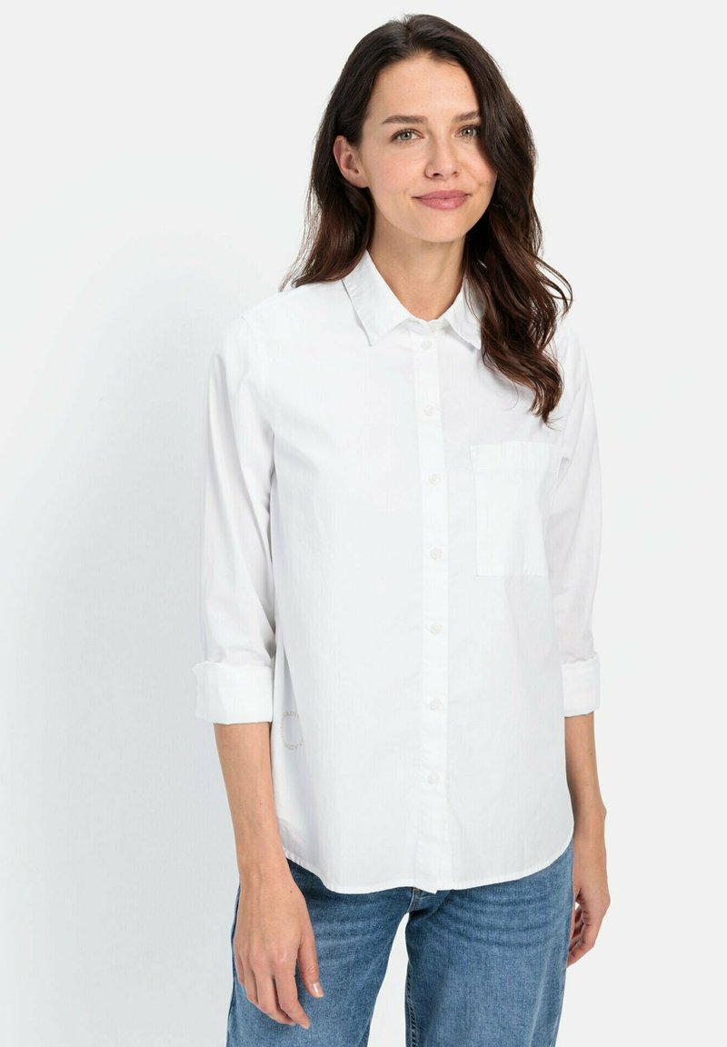 Chemise blanche à boutons avec une poche, manches longues roulées jusqu'au milieu de l'avant-bras, fabriquée en tissu léger, portée avec un jean bleu.