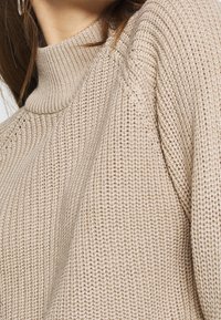 Beiger, gestrickter Pullover mit hohem Kragen, der ein strukturiertes Rippmuster und gesenkte Schultern aufweist. Der Stoff wirkt dick und warm.