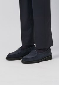 Navy blue suede loafers with a rounded toe and visible stitching, paired with dark trousers and dark socks, set against a light background.