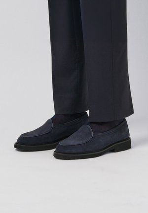 Mocassins en daim bleu marine avec un bout arrondi et des coutures visibles, associés à un pantalon foncé et des chaussettes foncées, sur un fond clair.