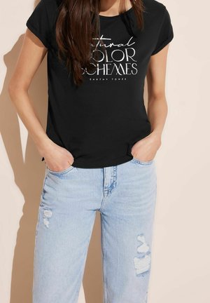 Frau trägt ein schwarzes T-Shirt mit dem Schriftzug "Natural Color Schemes Earthy Tones" und hellblaue, zerrissene Jeans, Hände in den Taschen, vor beigem Hintergrund.