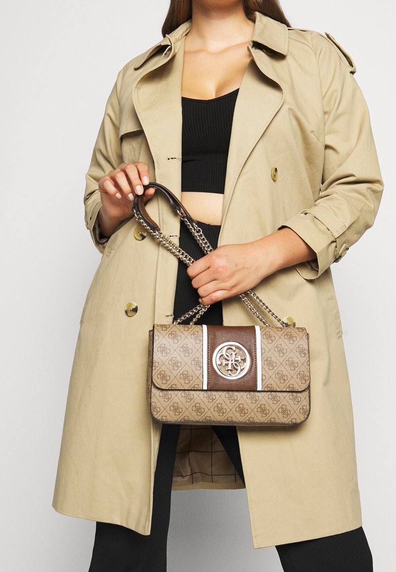 Donna con un trench beige e un outfit nero che tiene in mano una borsa di design marrone con tracolle a catena e dettagli del logo sul davanti.