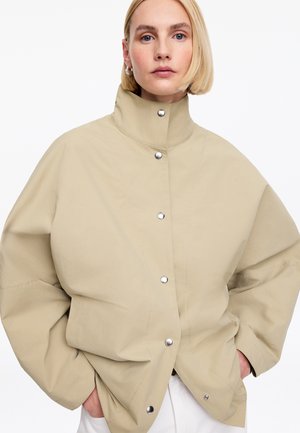 Frau mit kurzen blonden Haaren, die eine beige übergroße Jacke mit Druckknöpfen und hohem Kragen trägt, Hände in den Taschen, neutraler Hintergrund.