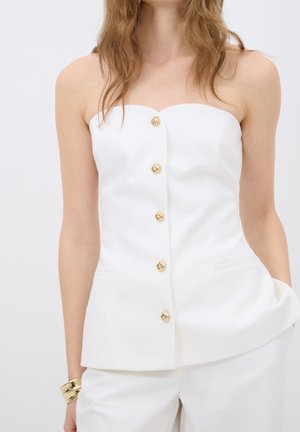 Trägerloses, weißes, maßgeschneidertes Oberteil mit goldenen Knöpfen vorne, kombiniert mit passenden weißen Hosen und einem massiven goldenen Armband am linken Handgelenk.