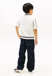 Παιδί όρθιο, φοράει λευκό πουλόβερ με κοντά μανίκια και μαύρες ρίγες, σκούρα τζιν με φαρδιά πόδια και ραμμένες τσέπες, και αθλητικά παπούτσια με κόκκινες και λευκές ρίγες.