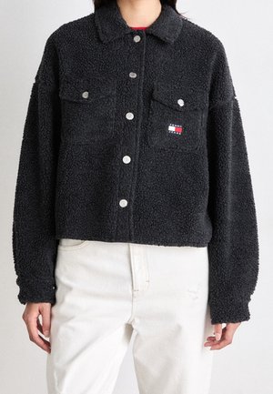 Czarna teksturowana kurtka zapinana na guziki z dwoma kieszeniami na piersi, noszona z kremowymi spodniami, z małym logo Tommy Jeans na lewej kieszeni.