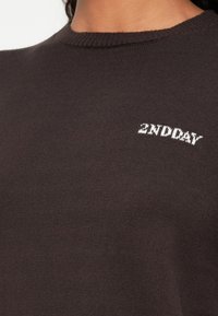 Detailní záběr na tmavohnědý pletený svetr s bílým vyšívaným nápisem "2ND DAY" poblíž horní části hrudníku.