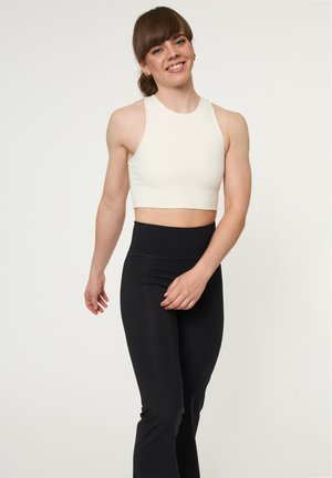 Naeratav inimene, kellel on pruunid juuksed seotud taga, kannab valget varrukateta crop-toppi ja kõrge vöökohaga musti retuuse, seistes ühevärvilise tausta ees.