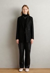 Zwarte pinstripe blazer over een zwart coltrui, gecombineerd met een zwarte nepleer broek. Het model draagt lichtgekleurde schoenen en minimale accessoires.