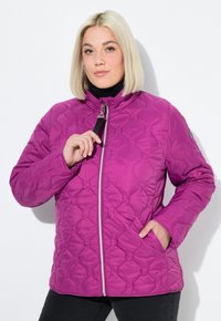 Blondýnka v jasně fialové prošívané bundě, která ji zapíná jednou rukou a druhou má v kapse, na bílém pozadí.