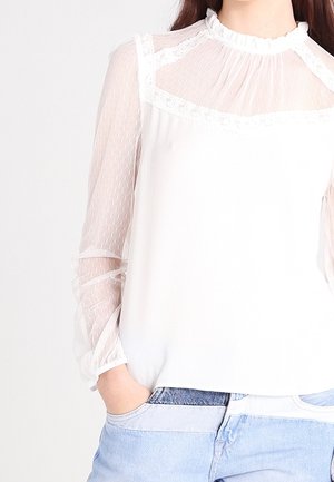 Biała bluzka z przezroczystymi, długimi rękawami w kropki i koronkowym obszyciem, zestawiona z jasnoniebieskimi dżinsami, ręka swobodnie włożona do kieszeni.
