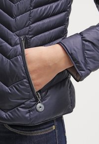 Veste rembourrée bleue marine présentant un motif matelassé en chevrons, une poche latérale zippée et une texture lisse et brillante.