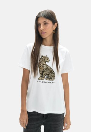 Mujer joven con cabello largo y oscuro que lleva pendientes geométricos dorados y una camiseta blanca con un gráfico de leopardo dorado y texto "LOLA CASADEMUNT".