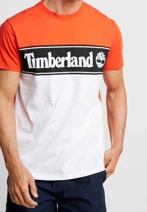 Homme portant un t-shirt Timberland orange et blanc à manches courtes avec logo et un pantalon bleu marine, les mains détendues sur les côtés.