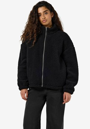 Jeune femme aux longs cheveux bouclés portant une veste en polaire sherpa noire zippée et un pantalon noir, debout devant un fond blanc.