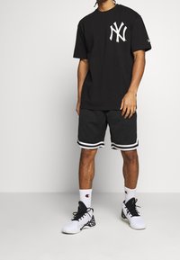 Man som bär en svart New York Yankees t-shirt, svarta sportshorts med vita ränder, vita Champion strumpor och vita och svarta sneakers.