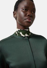 Dunkelgrüne Satinbluse mit hohem Ausschnitt, mit einem gemusterten Kragen in Grün und Off-White sowie einem Knopfverschluss am Hals.