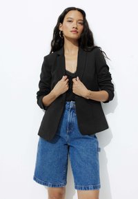 Blazer noir ajusté avec un col cranté, porté sur un débardeur en dentelle, associé à un short Bermuda en denim bleu. Texture de tissu lisse.