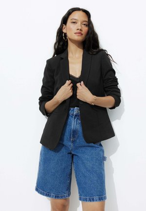 Blazer noir ajusté avec un col cranté, porté sur un débardeur en dentelle, associé à un short Bermuda en denim bleu. Texture de tissu lisse.