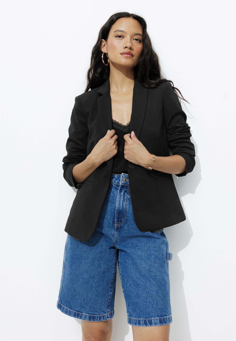Blazer noir ajusté avec un col cranté, porté sur un débardeur en dentelle, associé à un short Bermuda en denim bleu. Texture de tissu lisse.