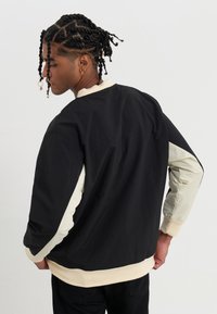 Chaqueta a color bloqueado en negro y crema con mangas largas, cuello y puños acanalados, fabricada con un tejido ligero que presenta una textura suave y un ajuste relajado.