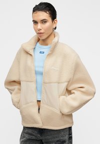 Veste en polaire beige avec un col haut, une fermeture éclair et des poches avant. Présente un panneau contrastant et un détail de logo subtil sur la poitrine.