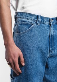 Jeans de mezclilla azul desgastado con corte recto, diseño de cinco bolsillos y cierre de botón de latón, mostrados con una mano descansando sobre el muslo.