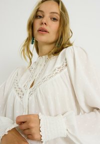 Blusa blanca con lunares texturizados, inserciones de encaje y puños con volantes. Cuenta con una tapeta de botones y una silueta fluida.