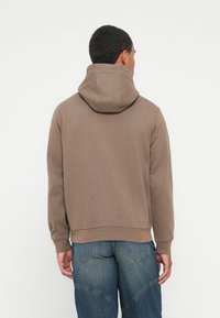 Hombre con una sudadera con capucha marrón y jeans de mezclilla azules, de pie de espaldas a la cámara frente a un fondo blanco liso.
