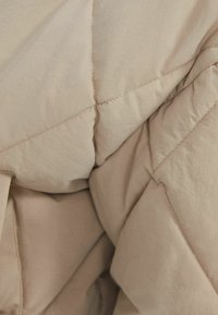 Beige kviltet dyne med en glat, mat tekstur. Har diamantsting og en subtil glans i stoffet. Blød og lagdelt udseende.