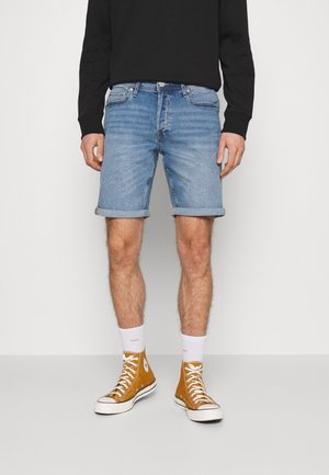 Uomo con pantaloncini di jeans blu risvoltati, calzini bianchi, sneakers alte giallo senape e una maglia nera a maniche lunghe, in piedi su un pavimento grigio.