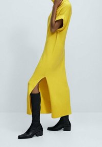 Keltainen neulemekko lyhyillä hihoilla ja sivuaukkoilla, jossa on ribbireunus. Yhdistetty mustiin polvipitkiin saappaisiin, joissa on paksu korko.