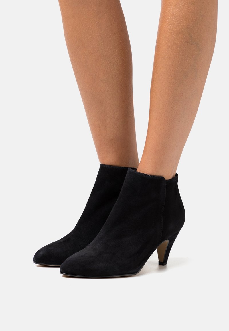 Ankelhöga svarta mockastövlar med spetsiga tår och låga avsmalnande klackar, bärs på bara ben mot en enfärgad bakgrund.