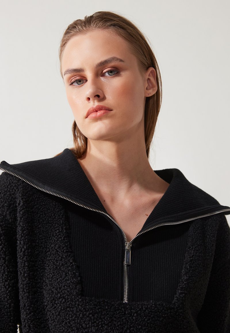 Vêtement noir à fermeture éclair en tissu texturé et duveteux, avec un col côtelé. Accent en métal argenté notable sur la fermeture éclair.