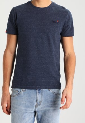 Hombre con una camiseta lisa azul oscuro de manga corta y vaqueros azul claro, de pie frente a un fondo liso y claro.