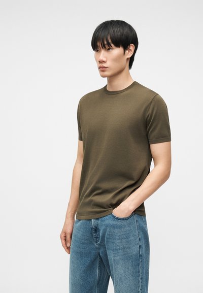 Νέος άνδρας με μαύρα μαλλιά, που φοράει πουκάμισο κοντομάνικο ελιάς και μπλε τζιν, στέκεται με το ένα χέρι στην τσέπη μπροστά σε απλό φόντο.