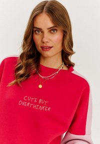Červená mikina se růžovými rukávy, s bílým vyšitým nápisem "CUTE BUT OVERTHINKER". Hladký materiál, uvolněný střih.