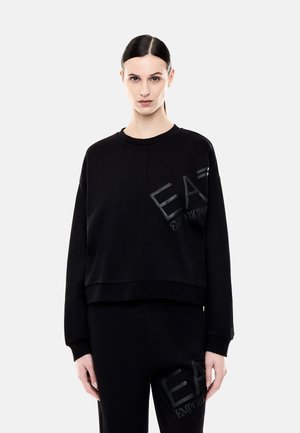 Femme aux cheveux foncés portant un sweat noir EA7 Emporio Armani et un pantalon assorti, face à un fond clair uni.