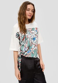 Korte mouwen t-shirt met een groene patroon aan de voorkant en een witte achterkant, met een strikdetail bij de taille en glanzende zwarte broek.