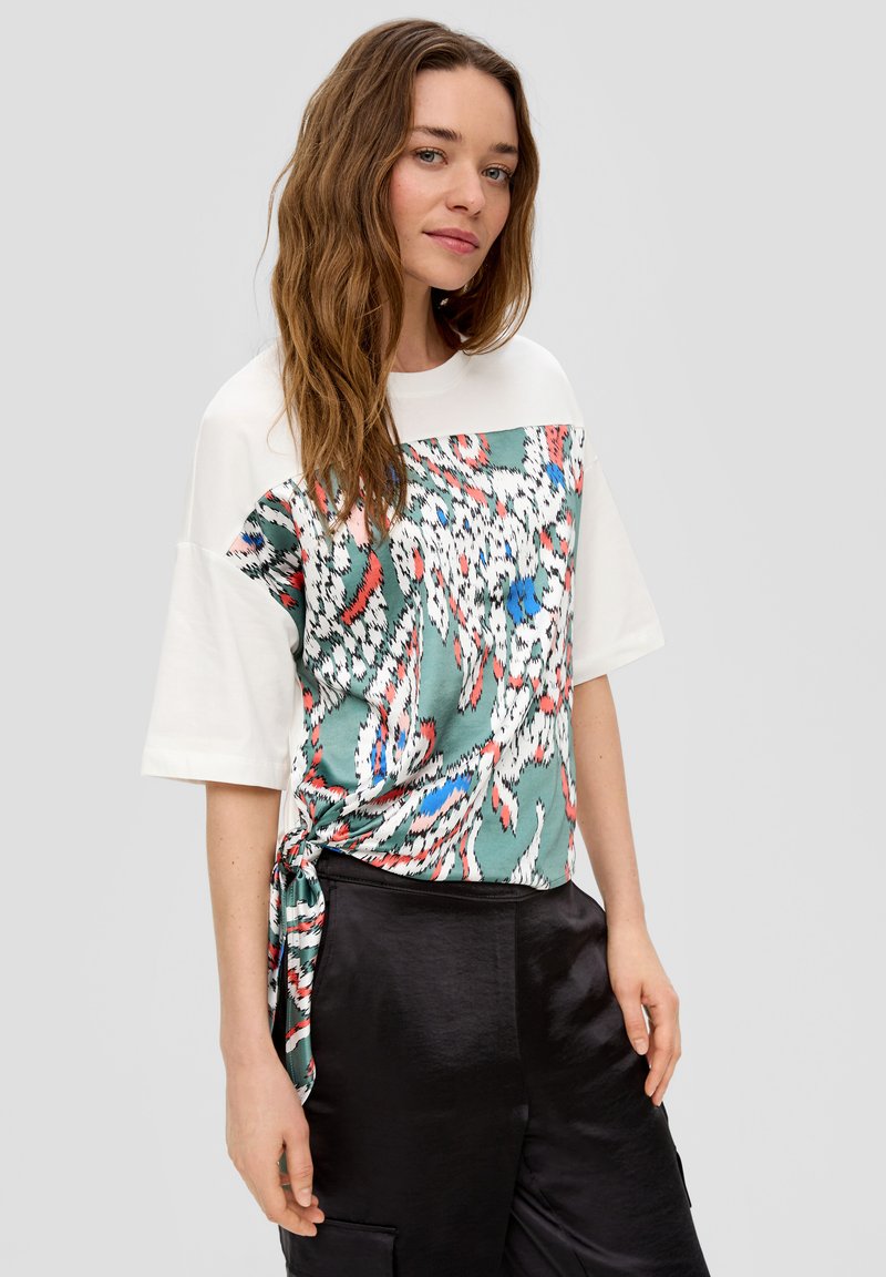 Korte mouwen t-shirt met een groene patroon aan de voorkant en een witte achterkant, met een strikdetail bij de taille en glanzende zwarte broek.