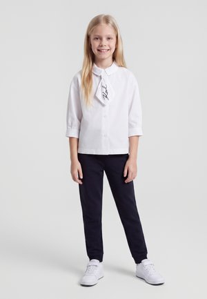 Smilende pige med langt blond hår iført hvid skjorte med krave, sorte bukser og hvide sneakers, står foran en ensfarvet baggrund.